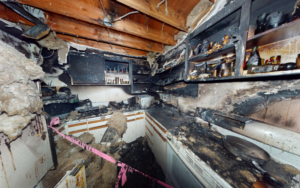 Fire-damaged kitchen with heavy soot and smoke damage on cabinets, countertops, and walls, exposed ceiling beams, burned shelving, and debris from insulation and firefighting efforts.